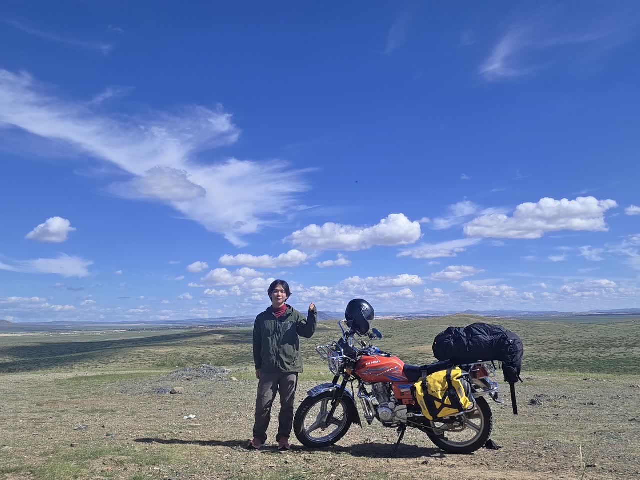丘の上で、荷物を積んだ赤いバイクの横に立つ筆者。遠くまで草原が続く